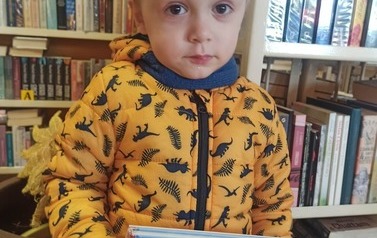 Chlopiec o blond wlosach i w ż&oacute;łtej kurtce trzyma książkę z wyprawki czytelniczej.