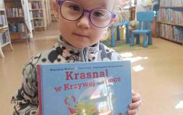 Dziewczynka o blond włosach i w okularkach pokazuje swoją nową książkę z wyprawki czytelniczej.