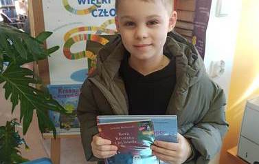 Chłopiec stoi przy tablicy promującej akcję Mała Książka Wielki Człowiek i trzyma książkę wypożyczoną i otrzymaną z wyprawki czytelniczej.