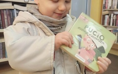 Chłopiec o blond włosach i w jasnej kurteczce patrzy na swoją wypożyczoną książeczkę.