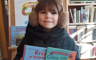 Dziewczynka stoi przy tablicy promującej akcję Małego Czytalnika i trzyma książkę wypożyczoną i otrzymaną z wyprawki czytelniczej.