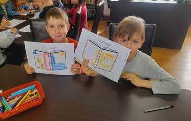 Dw&oacute;ch chlopc&oacute;w przy stole pokazuje swoje projekty okładki,  w tle widać z tyłu inne dzieci.