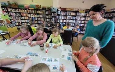 Dzieci siedzą przy stołach i wyklejają obrazkami historyjkę o powstaniu ksiązki. Przy jednym stoliku stoi bibliotekarka i patrzy na ich pracę.