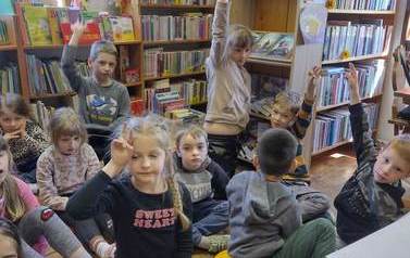 Dzieci siedzą na siedziskach i słuchają bibliotekarza. Niekt&oacute;re z dzieci podnoszą rękę zgłaszając się.