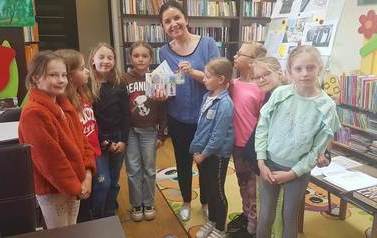 Bibliotekarka stoi z paroma dziewczynkami trzymając od nich laurki i świeczkę. Bardzo się uśmiecha.
