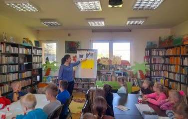 Dzieci siedzą przy stołach i patrzą do przodu na bibliotekarkę, kt&oacute;ra przyczepia do metalowej tablicy ilustrację dotyczącą powstawania książki.