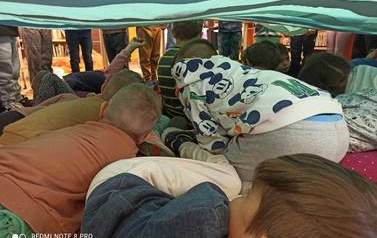 Pod dużą rozciągniętą przez dzieci matą kucają dzieci.