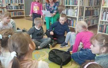 Dzieci siedzą na dywanie, a Pani wychowawczyni czyta im z kartki wiersz o bibliotece i bibliotekarce.
