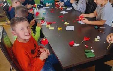 Dzieci przy stołach robią z zielonej bibuły łodygi i listki do kwiat&oacute;w.