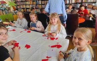 Przy przednim stole siedzą dziewczynki i z uśmiechem wycinają z czerwonej bibuły płatki do kwiata, z tyłu widać stolik z chłopcami.