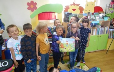 Dzieci stoją na dywanie i trzymają otwartą książkę, a przed nimi na podłodze leży uśmiechnięty chłopiec.