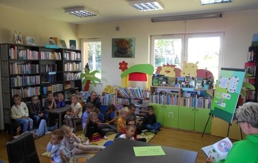 Dzieci siedzą na dywanie i słuchają bajki, kt&oacute;rą czyta im zaproszony gość.