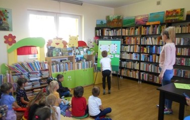 Dzieci siedzą na dywanie, a z przodu przy metalowej tablicy stoi chłopiec i wykonuje na niej zadanie. Z boku stoi bibliotekarka.
