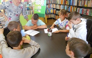Chłopcy siedzą przy stole i wykonują kartę pracy. Przy ich stole stoi bibliotekarka i patrzy.