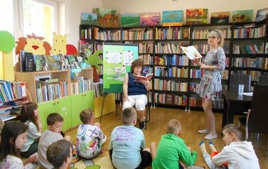 Dzieci siedzą na dywanie,a z przodu siedzi gośc i czyta im bajkę. Z prawej strony stoi z otwartą książką bibliotekarka.