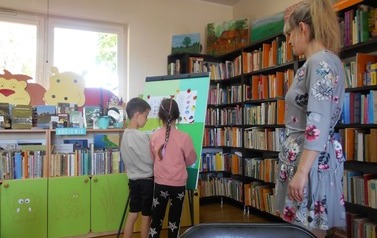 Chłopiec z dziewczynką stoją przy tablicy i wykonują zadanie. Obok stoi bibliotekarka i na nich patrzy.