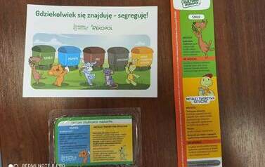 Na stole leżą: poczt&oacute;wka, zakładka do książek i magnes o segregacji śmieci.