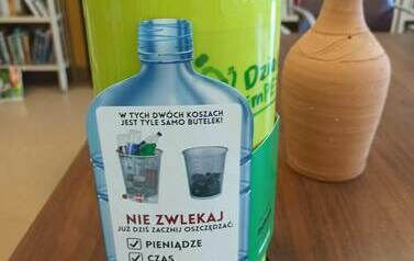 Na stole postawiona jest zielona zgniatarka do butelek z naklejoną etykietą butelki. 