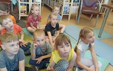 Dzieci siedzą na dywanie i karimatach i słuchają czytanej bajki.