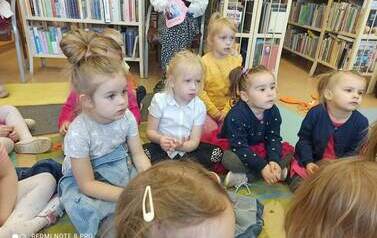 Dzieci siedzą na dywanie i słuchaja bajki, z tyłu widać na krześle opiekuna dzieci.