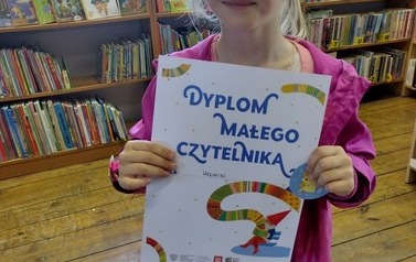Dziewczynka o blond włosach i w r&oacute;żowej kurtce pokazuje dyplom i w ręku trzyma naklejkę "super czytelnika".