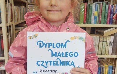 Dziewczynka w r&oacute;żowej kurtce trzyma dyplom, kredki i naklejkę. Nad oczami ma namalowane rzęsy w kształcie tr&oacute;jkąta.