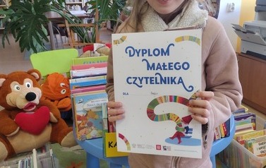 Dziewczynka o blond włosach pokazuje dyplom, w tle widać książki  i misie na dywanie.