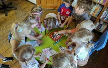 Dzieci stoją przy okrągłym stoliku i wykonują pracę plastyczną - loda z papieru i kolorują go kredkami.