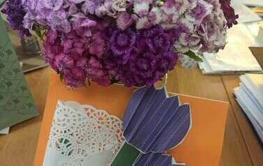 Na biurku leżą żywe kwiaty fioletowe, a obok na białej serwetce leżą własnoręcznie zrobione fioletowe kwiatki z papieru.