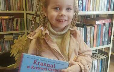 Uśmiechnięta dziewczynka o blond włosach trzyma w ręku swoją wypożyczoną książkę i książkę z wyprawki czytelniczej.