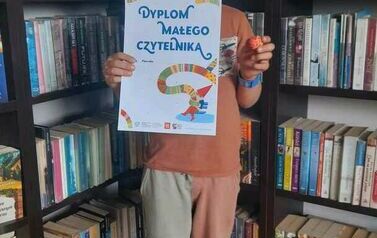 Chłopiec stoi w bibliotece w Pinczynie trzymajć sw&oacute;j dyplom. W prawej ręce trzyma małą piłeczkę. 