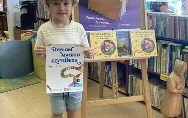 Chłopiec w kapeluszu pokazuje w bibliotece w Zblewie sw&oacute;j dyplom. Za nim stoi stojak z plakatem i książkami dotyczącymi akcji.