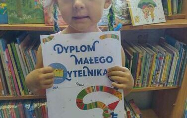 Dziewczynka o blond włosach w dw&oacute;ch warkoczykach pokazuje w bibliotece w Borzechowie sw&oacute;j dyplom. 