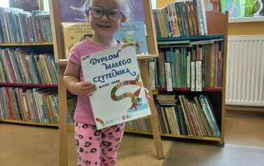 Mała dziewczynka w okularkach uśmiecha się trzymając dypolm Małego Czytelnika.Za nią stoi stojak z plakatem dotyczącym akcji MKWC. 