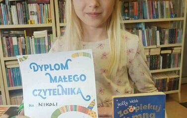 Dziewczynka o blond włosach i w kapeluszu trzyma w prawej ręce dyplom,a w lewej wypożyczoną książeczkę.