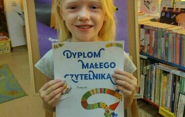 Dziewczynka w blond wlosach pokazuje w bibliotece w Zblewie sw&oacute;j dyplom Małego Czytelnika. Za Nią stoi stojak z plaktem dotyczącym projektu Mała Książka Wielki Człowiek.