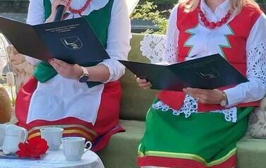 Dwie Panie w strojach kociewskich siedzą przy stoliku z nakryciem kawowym. Jedna z nich czyta tekst " Nad Niemnem". 