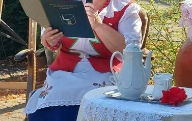 Pani z KGW siedzi przy stoliku z nakryciem kawowym  i czyta tekst o Janie i Cecylii.