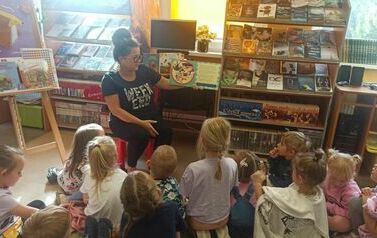 Dzieci siedzą na dywanie, a Pani z biblioteki czyta im bajkę o jesieni.