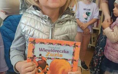 Dziewczynka w blond włosach trzyma książeczkę pt. "Wiewi&oacute;reczka Zuzia" pokazując ją.