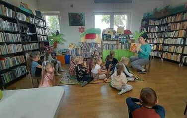 Dzieci siedzą na dywanie i patrzą do przodu w stronę bibliotekarki, kt&oacute;ra pokazuje im ilustrację z książki o wiwewi&oacute;rce.