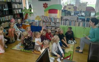 Dzieci siedzą na dywanie i małych krzesełkach i słuchają tego co m&oacute;wi z przodu siedząca bibliotekarka.