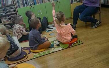 Dzieci siedzą na siedziskach i słuchają zagadek, kt&oacute;re czyta im bibliotekarka. Jedna dziewczynka podnosi rękę.