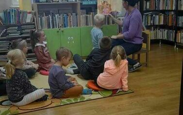Dzieci siedzą na dywanie i patrzą w ilustracje bajki, kt&oacute;rą czyta im bibliotekarka.