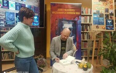 Pisarz podpisuje książkę, obok stoi czytelniczka w okularach.