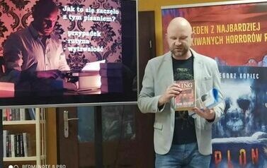 Pisarz pokazuje na dużym ekranie prezentację, za Nim stoi baner reklamujący książkę "Plon".