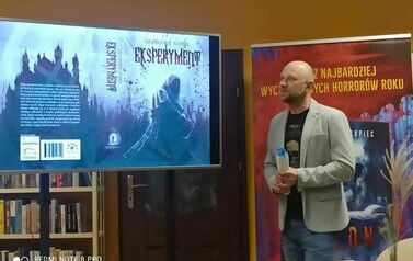 Pisarz pokazuje na dużym ekranie prezentację, za Nim stoi baner reklamujący książkę "Plon".