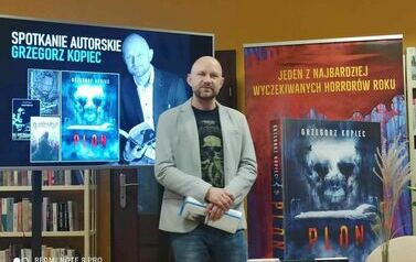 Pisarz stoi pomiędzy dużym telewizorem na kt&oacute;rym jest wyświtlana prezentacja pisarza, a banerem reklamującym książkę pisarza "Plon".