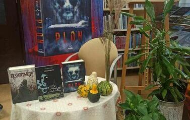 Stolik nakryty obrusem na kt&oacute;rym położone są ksiązki pisarza i małe dynie. Przy stoliku jest puste krzesło, obok kwiaty doniczkowe i z tyłu baner reklamujący książkę "Plon",a po prawej stronie tablica korkowa z informacjami o pisarzu. 