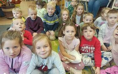 Dzieci siedzą na dywanie i słuchają bajki. Niekt&oacute;re z nich się ładnie uśmiechają.